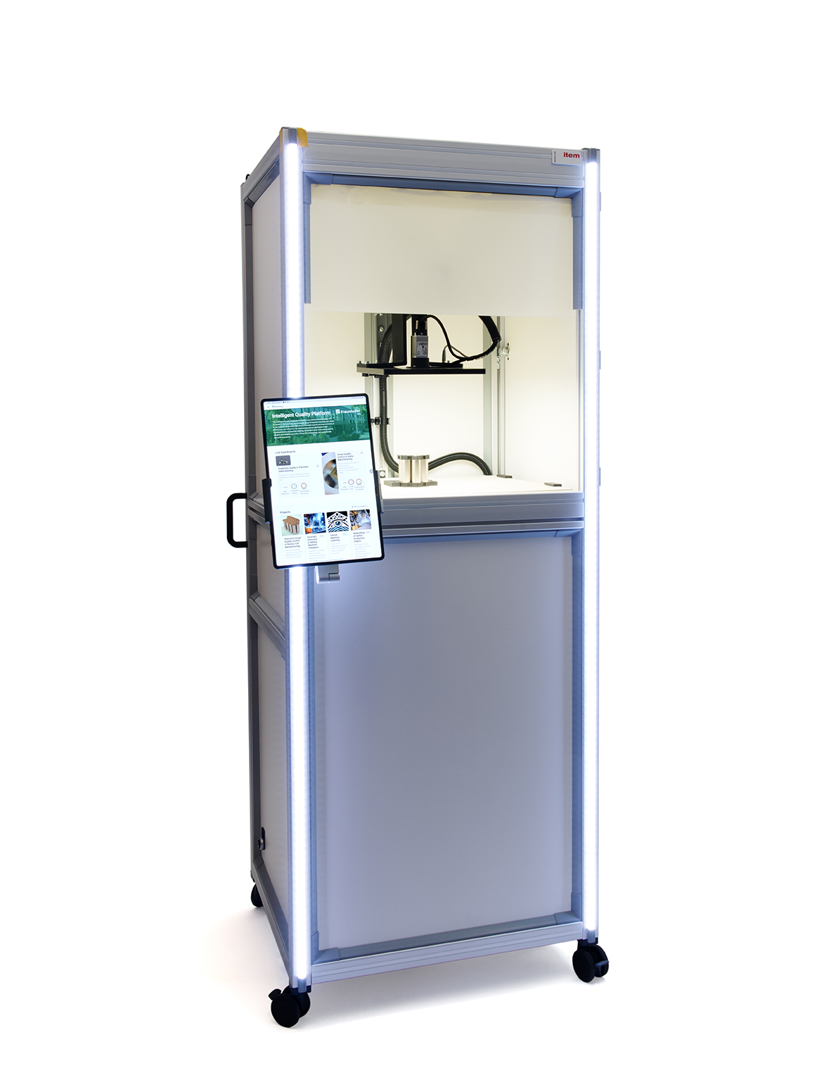 A tall, rectangular machine with a metallic frame and transparent glass front, featuring internal machinery visible through the glass. The machine is illuminated with white LED lights along the edges. To the left, a tablet stands on a holder, displaying information with a green and white interface. The machine is mounted on four black wheels for mobility, and the overall color scheme is predominantly silver and white. The background is bright and neutral, enhancing the visibility of the device.